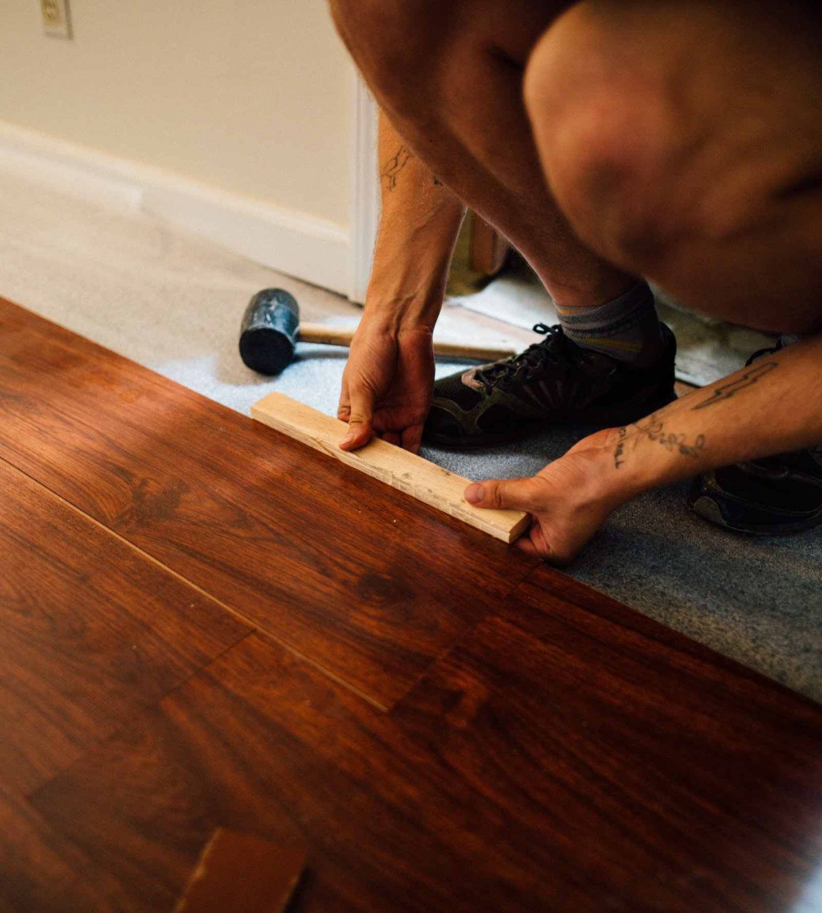Hands tapping a laminate floor plank into place with a wooden block, a rubber mallet beside them